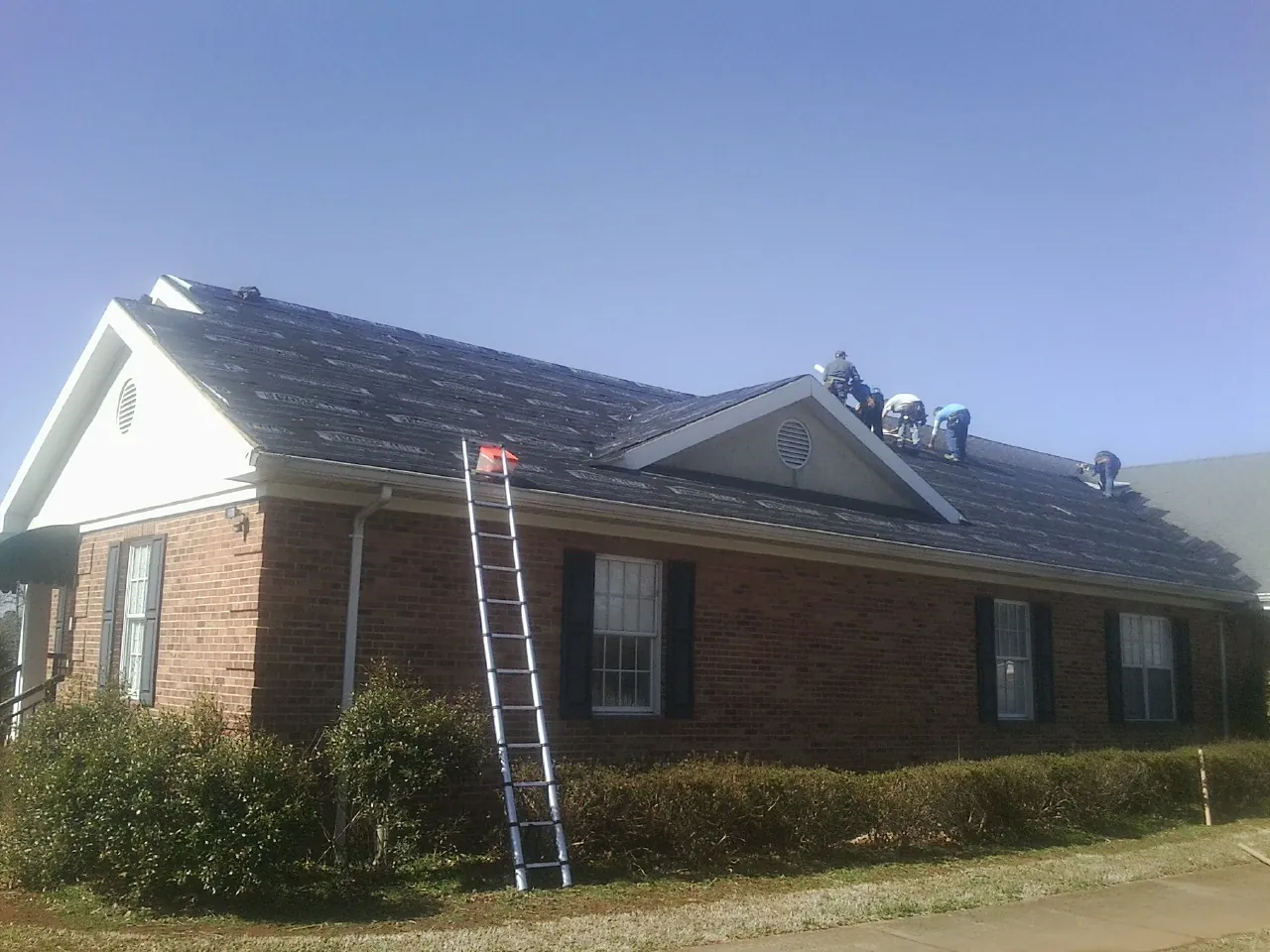 Crew tearing shingles off a brick ranch roof with a ladder set — Metal Roof Repair work by our crew in Glen Gardner