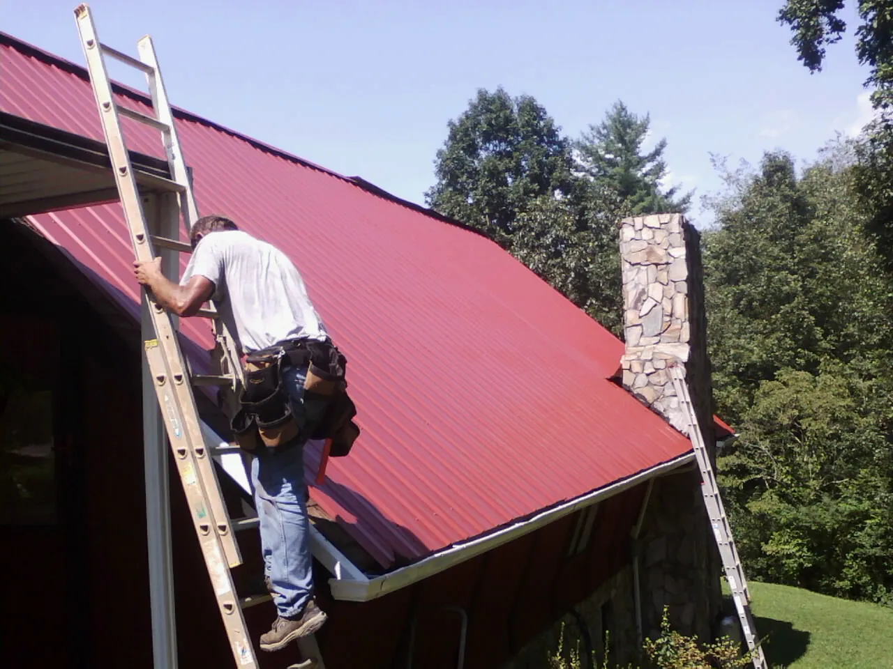 Roofer on a ladder beside a red metal roof and stone chimney — Soffit & Fascia Repair by Roofing Brothers in Glen Gardner